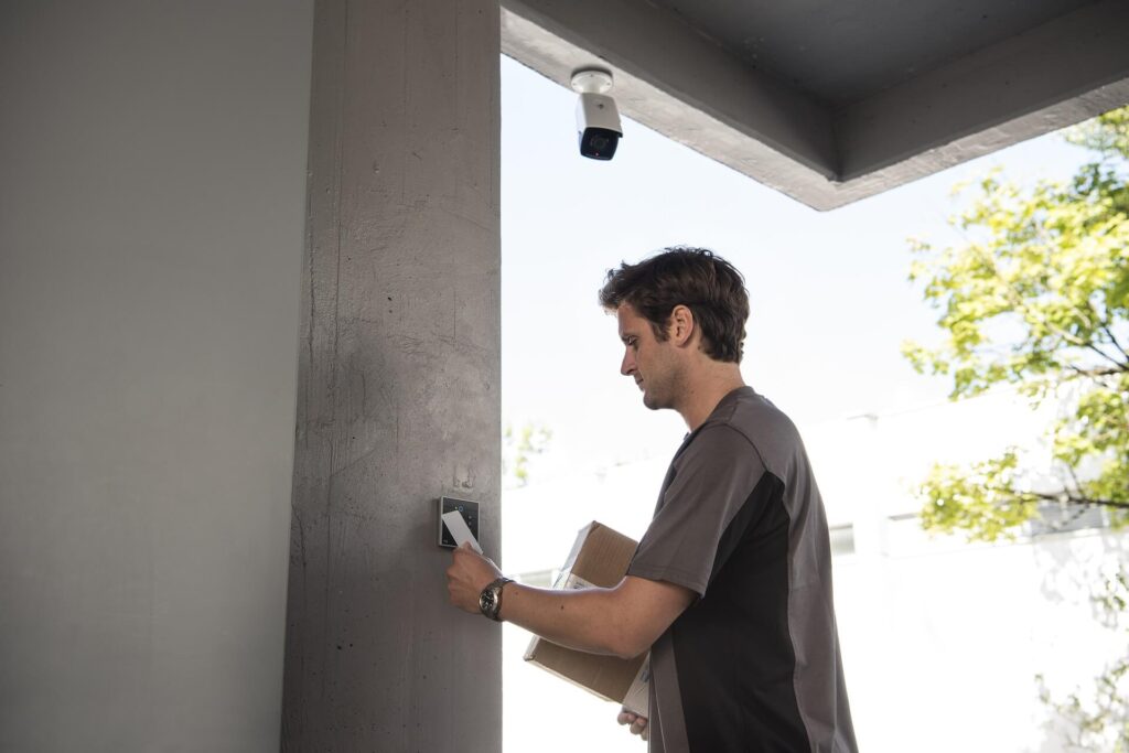 Um homem com uma camiseta cinza com uma faixa preta passando seu cartão num dispositivo na parede que detecta a sua entrada. Ao fundo vemos os galhos de uma árvore e acima do homem há uma câmera de segurança.