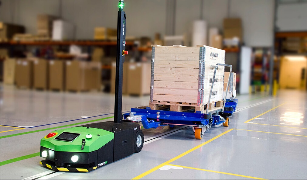 no canto inferior esquerdo possui um robô AGV de transporte na cor verde seguindo uma linha no chão, carregando materiais. No fundo da imagem, é possível ver um depósito de materiais de uma indústria